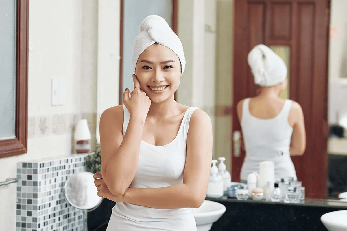 Asian woman in bathroom with white towel wrapped around head, face on hands and smiling