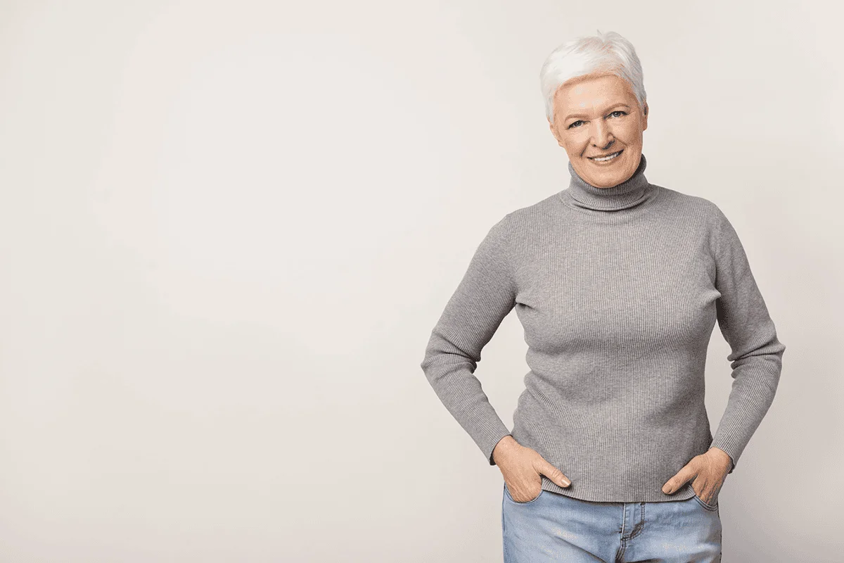 older woman with hands in pockets smiling on a toupe background